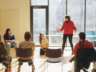 A woman in red stands next to two other women, one kneeling & one seated. She is directing a scene for the rest of the class who are sat on chairs facing her.
